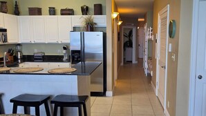 A kitchen with white cabinets, a stainless steel refrigerator, a microwave, and a coffee maker. There are three stools and a small island with a woven mat. - {L@@K}  Beach FRONT, Home @ the Beach w/Beach Chair Service {L@@K} (Panama City Beach)