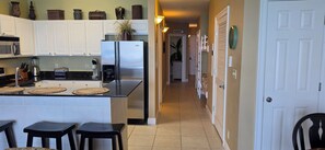 A kitchen with white cabinets, a stainless steel refrigerator, a microwave, and a coffee maker. There are three stools and a small island with a woven mat.