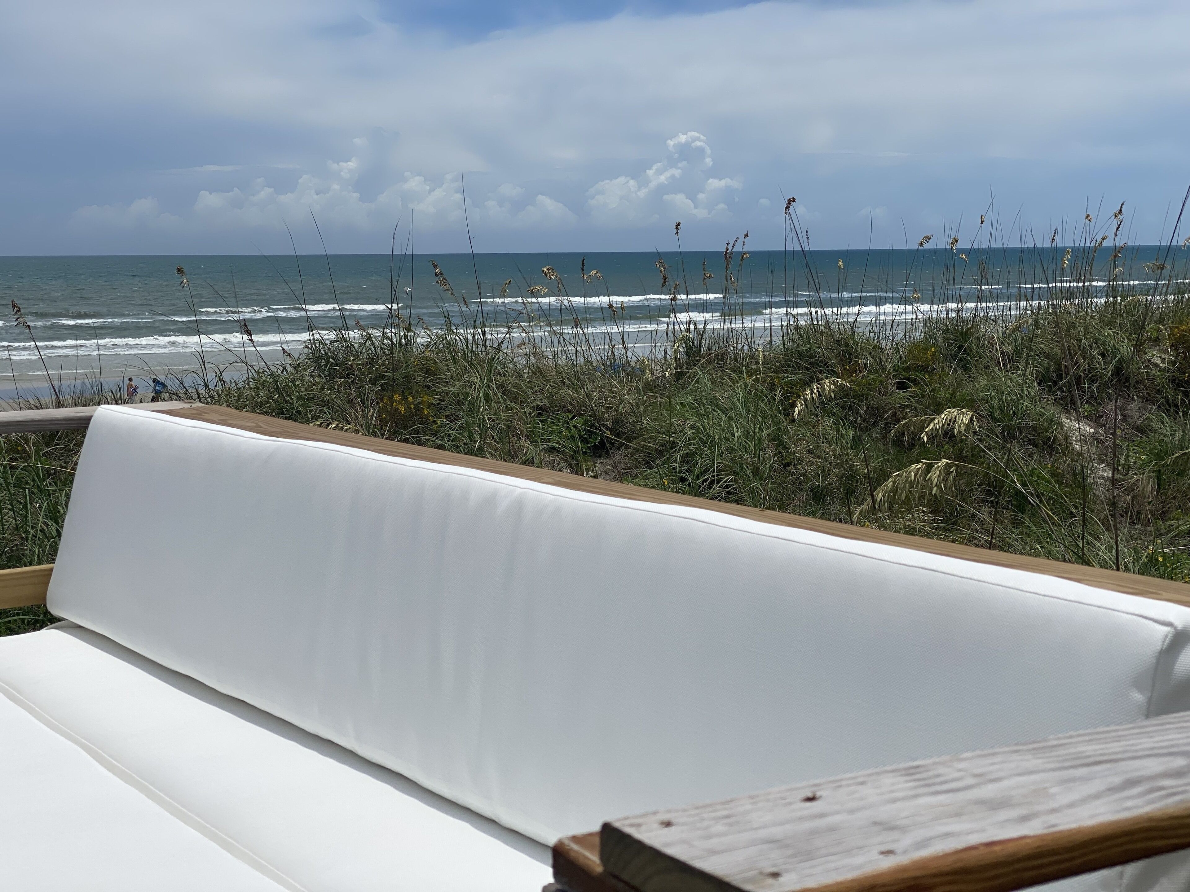 A white cushioned bench with a wooden frame, overlooking a beach with waves and seaweed.