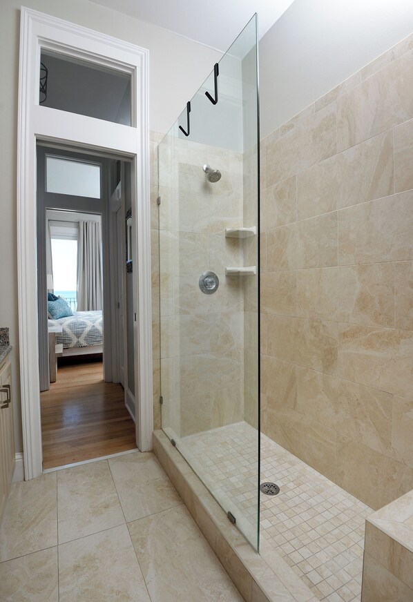 Un baño moderno con cabina de ducha de vidrio, azulejos color beige y una habitación visible con cama y cortinas.