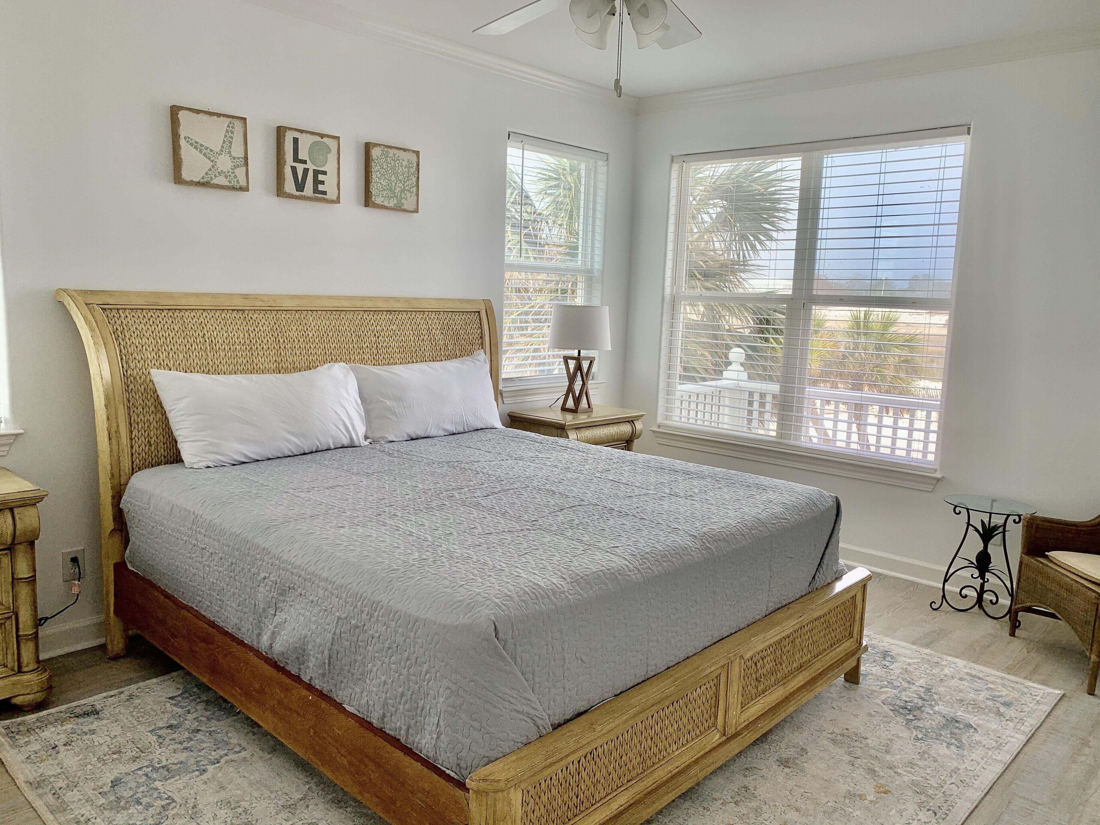 Un dormitorio con cama de madera, ropa de cama blanca, una mesita de noche con lámpara y vista a palmeras a través de la ventana.