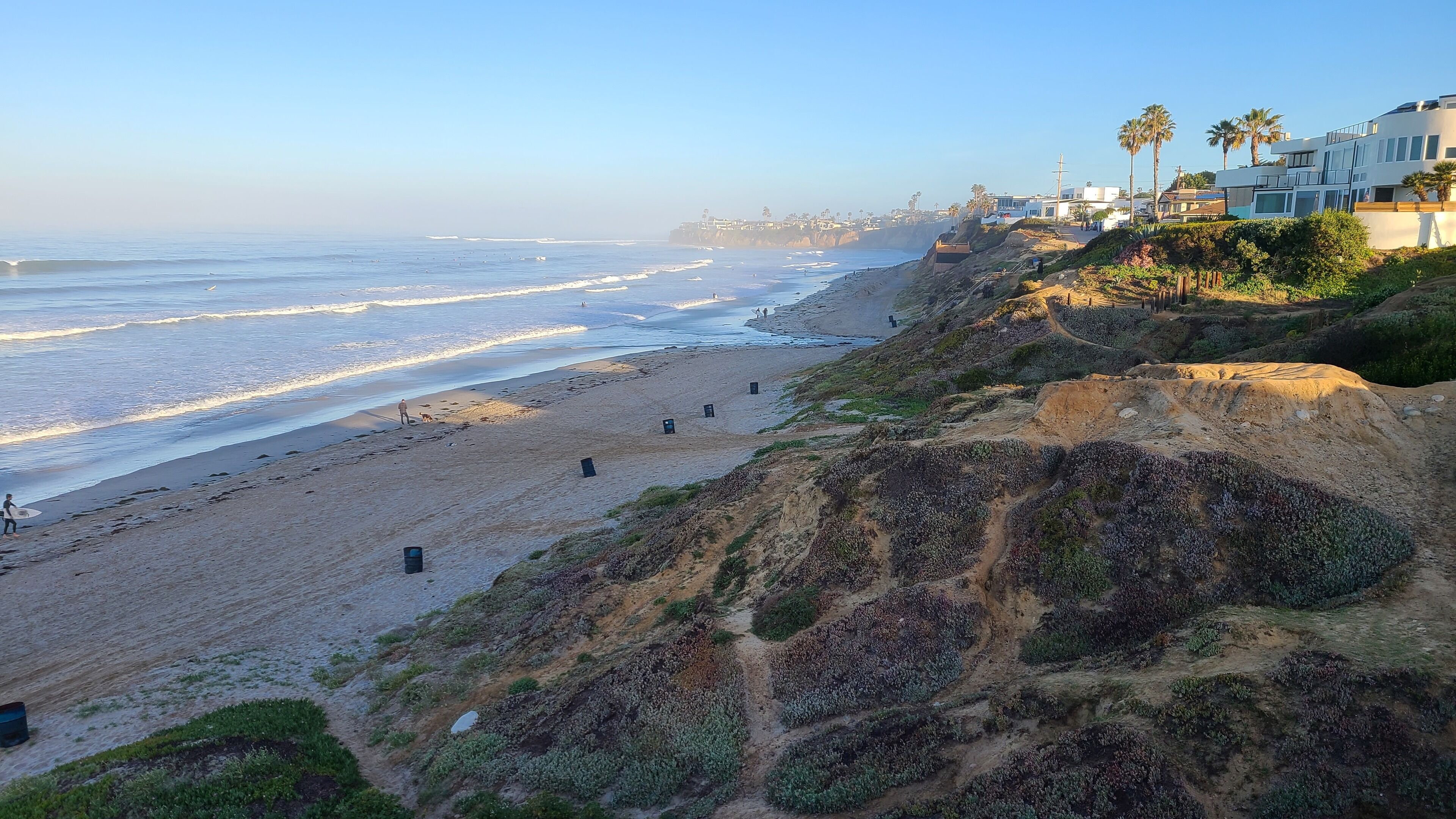 A coastal beach with a sandy shore, a rocky cliff, and modern buildings in the background.