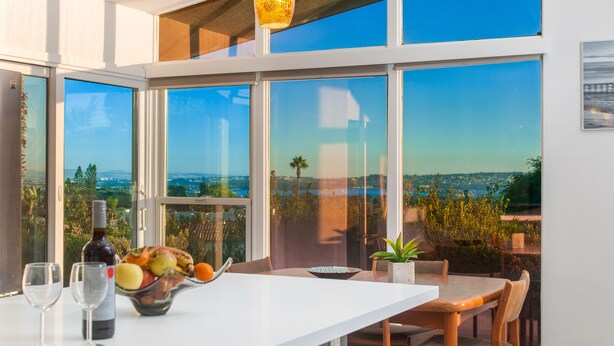 A dining area with a table, chairs, a bowl of fruit, wine glasses, and a bottle. Large windows offer a view of the landscape. - Newly Remodeled with VIEWS of Downtown Ocean and Bay! (San Diego)