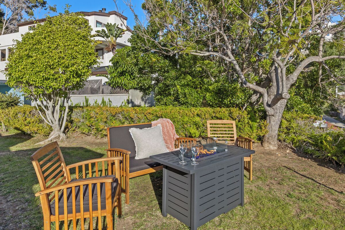 Un espacio al aire libre con mesa y sillas, rodeado de follaje y un árbol.