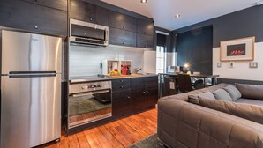 A modern kitchen with a refrigerator, oven, microwave, and dark cabinetry. There is a dining area with a table and chairs, and a bed with a gray headboard.
