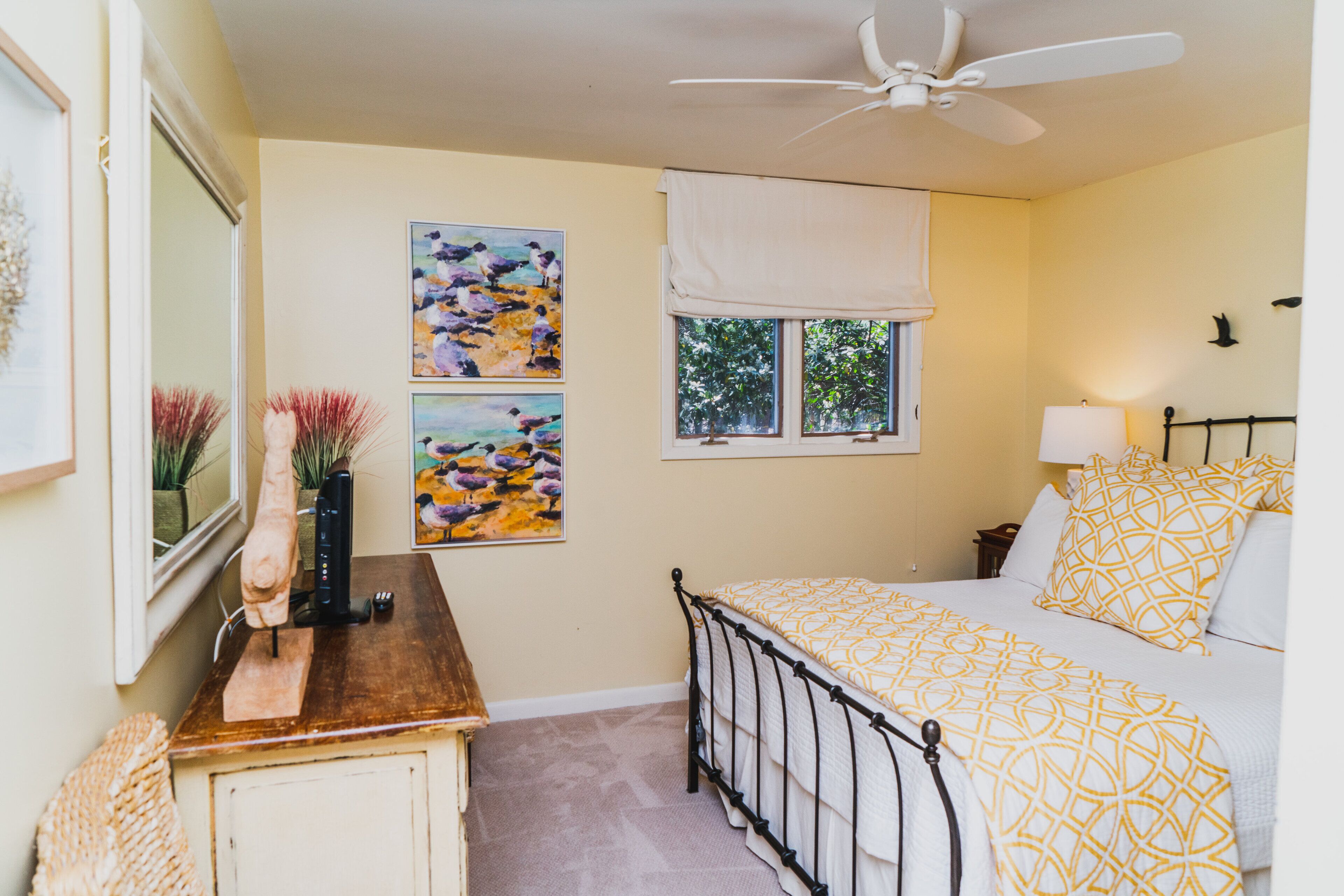 A bedroom with a bed, a ceiling fan, a window with a white blind, and two paintings on the wall.