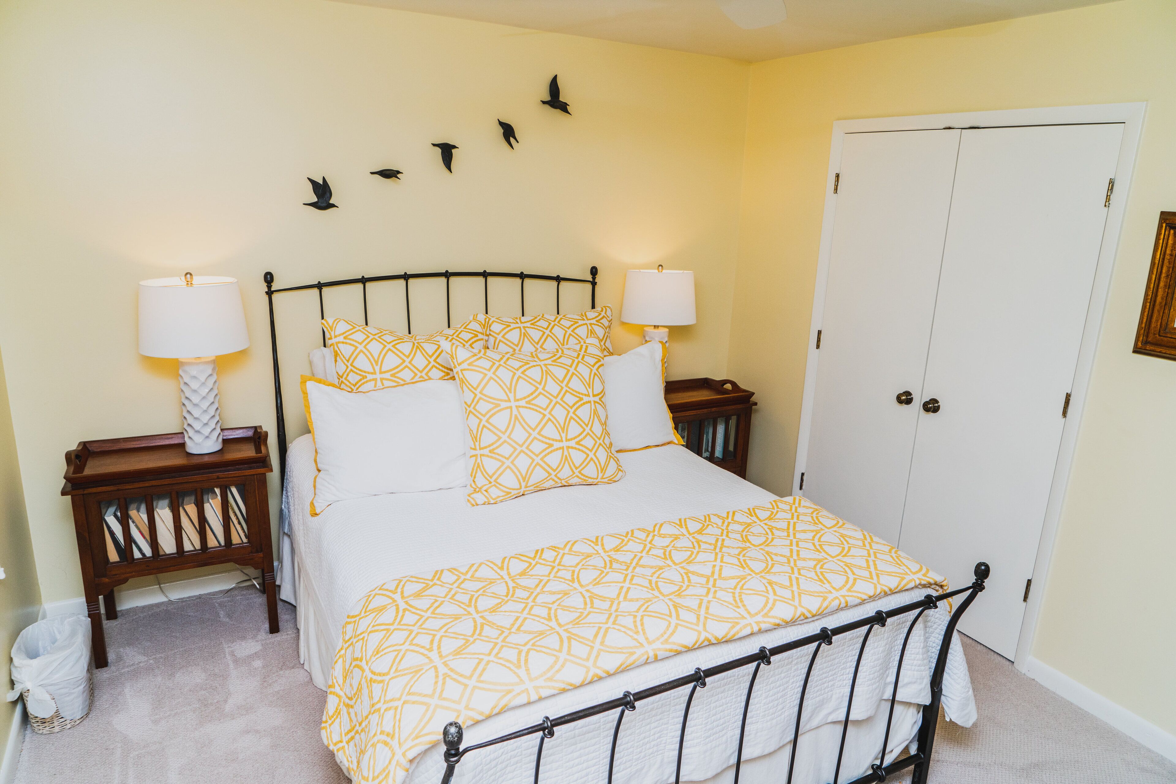 A bedroom with a bed, two bedside tables with lamps, a wooden nightstand, a white door, and wall decorations.