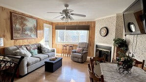 A living room with a sofa, armchairs, a coffee table, a dining table with chairs, a fireplace, a ceiling fan, and a large painting on the wall.