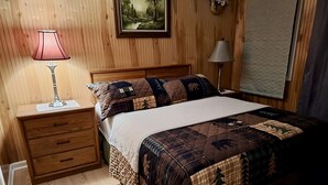 A neatly arranged bedroom with a wooden headboard, a quilted bedspread, a nightstand with a lamp, and a framed picture on the wall.