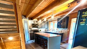 A modern kitchen with wooden beams, a central island, bar stools, and a refrigerator with magnets. - A large, newly remodeled 7,000 sq ft cabin, new hot tub, minutes from Deadwood! (Lead)