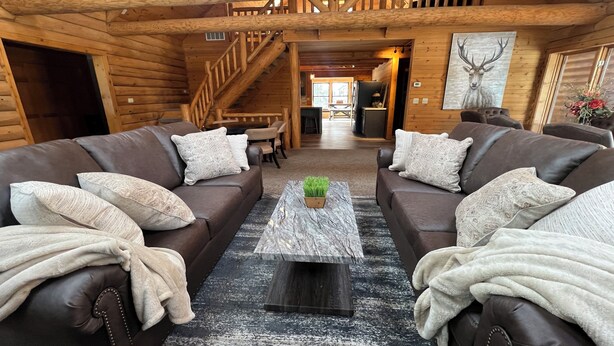 A log cabin interior with two leather sofas, a central coffee table, and a staircase leading to an upper level. - A large, newly remodeled 7,000 sq ft cabin, new hot tub, minutes from Deadwood! (Lead)