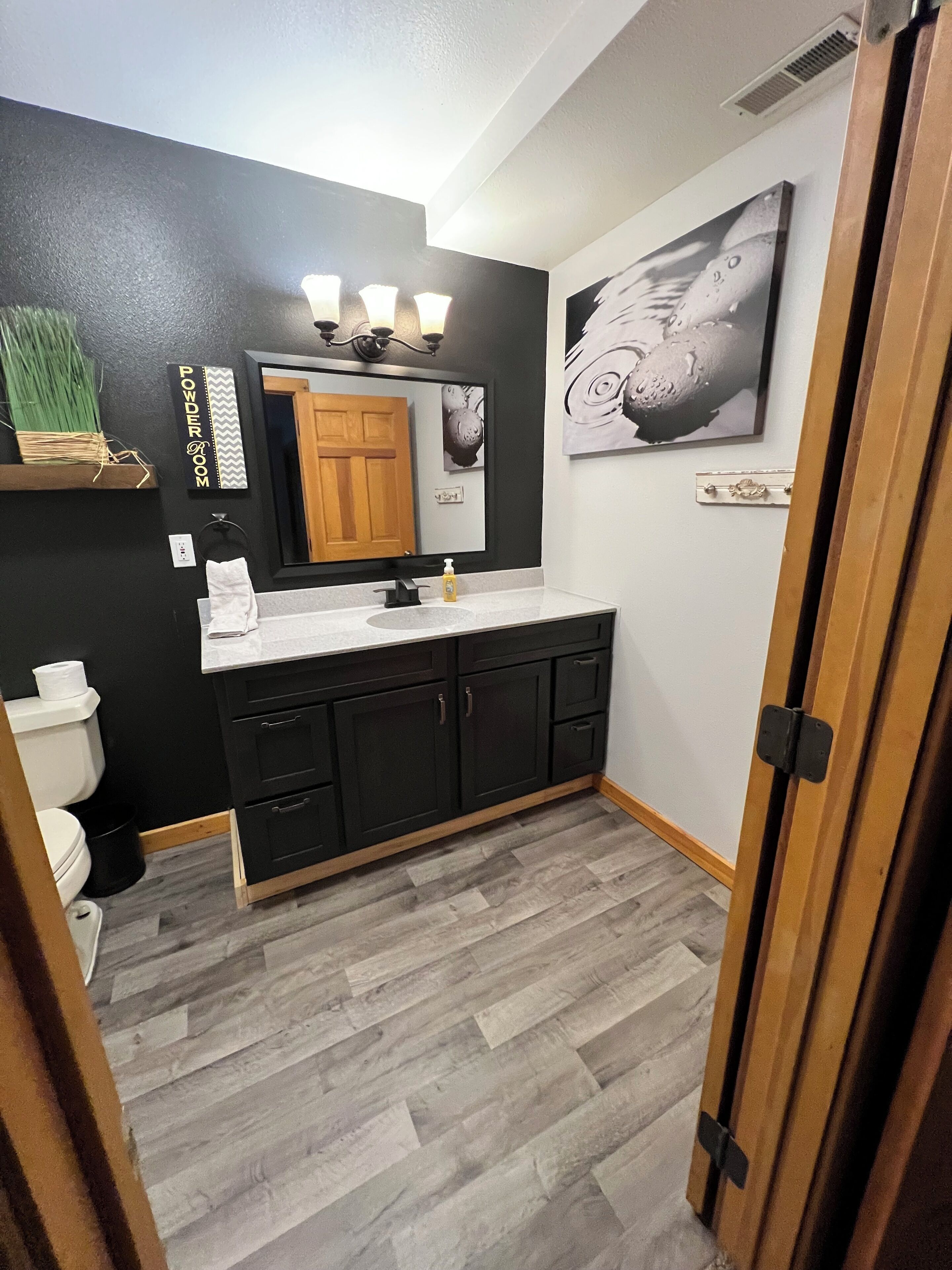 A bathroom with a black wall, a wooden door, a white toilet, a black cabinet, a mirror with lights, and a gray floor.