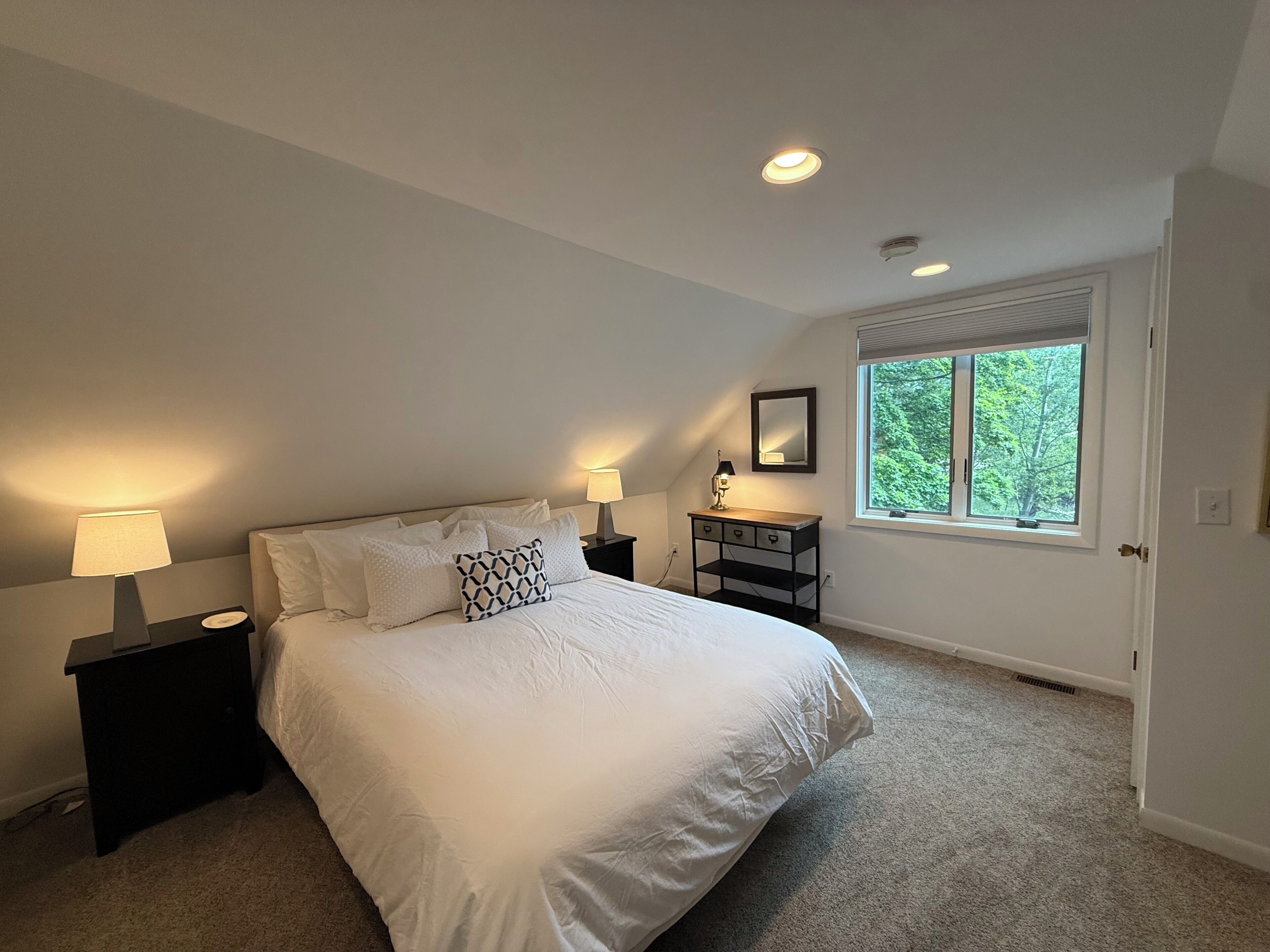 A bedroom with a bed, nightstands, a dresser, a window with a view of trees, and recessed ceiling lights.