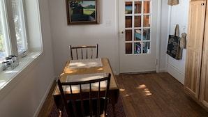 A dining area with a wooden table, chairs, a pendant light, a window with blinds, a door with a frosted glass panel, and a painting on the wall.
