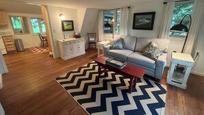 A modern living room with a grey sofa, a wooden coffee table, and a chevron-patterned rug. There is a kitchen area with wooden cabinets and a dining table.