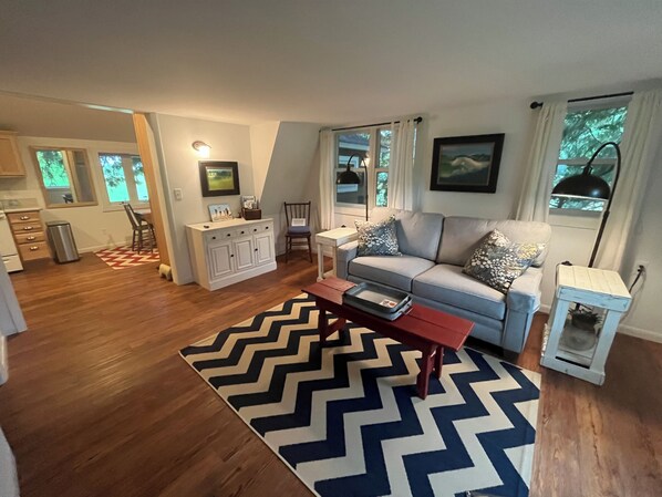 A modern living room with a grey sofa, a wooden coffee table, and a chevron-patterned rug. There is a kitchen area with wooden cabinets and a dining table.