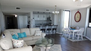 A living room with a beige sofa, a glass coffee table, and a patterned rug. There is a dining area with white chairs and a table.