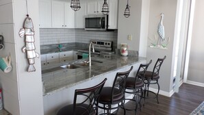 A kitchen with white cabinets, a granite countertop, a stainless steel sink, a microwave, and a stove. There are three bar stools and a decorative fish sculpture.