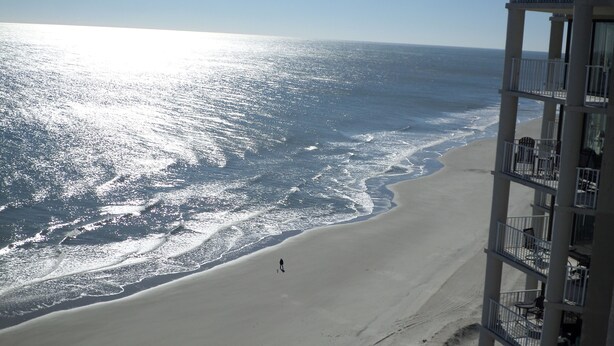 A beach with a person walking, a building with balconies, and the ocean.