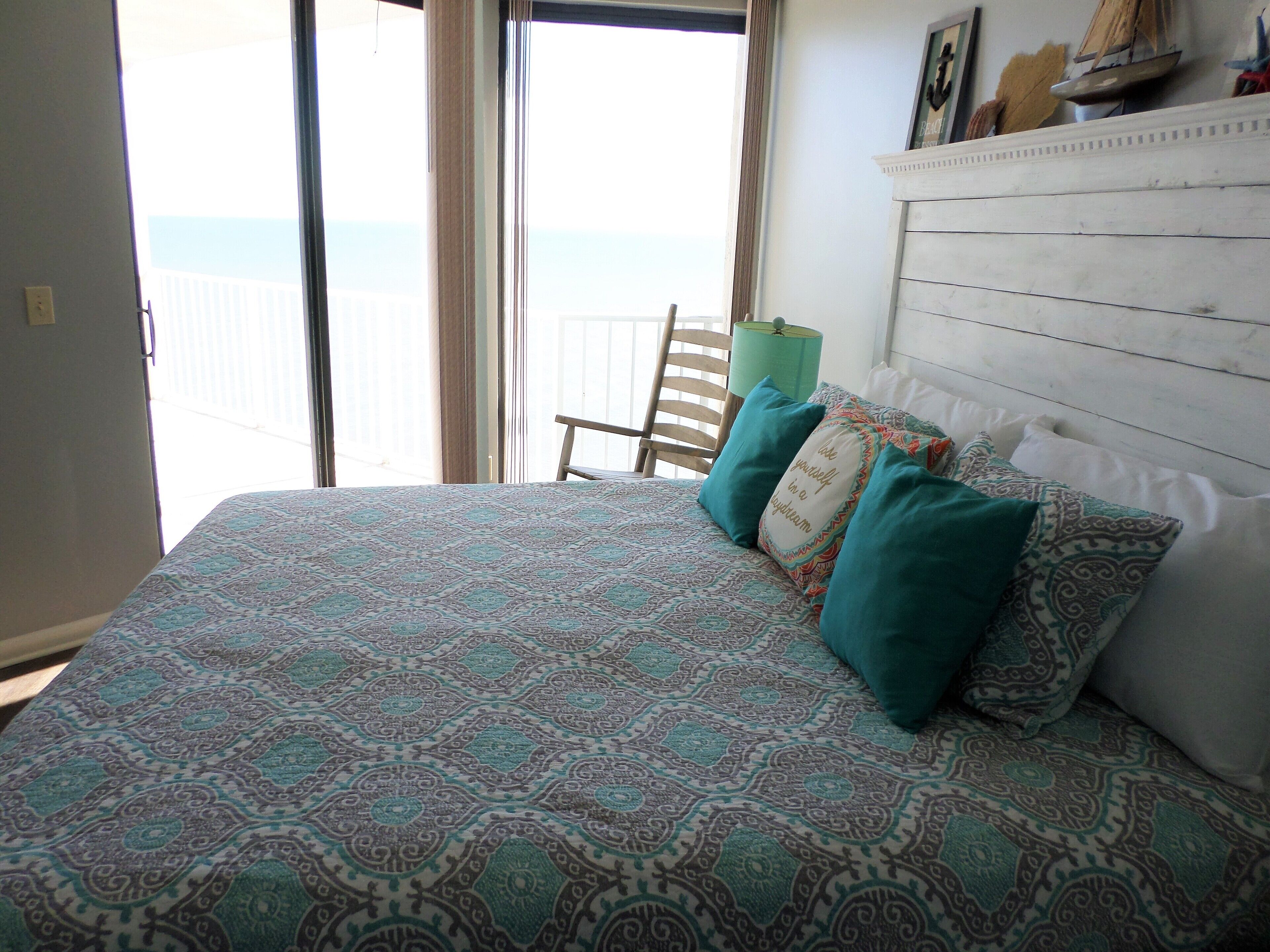 Un lit avec une literie à motifs, une tête de lit en bois et une vue sur la mer depuis la porte d’un balcon.
