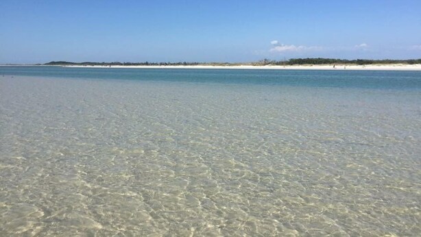 A clear beach with shallow water, distant sand dunes, and a clear sky.