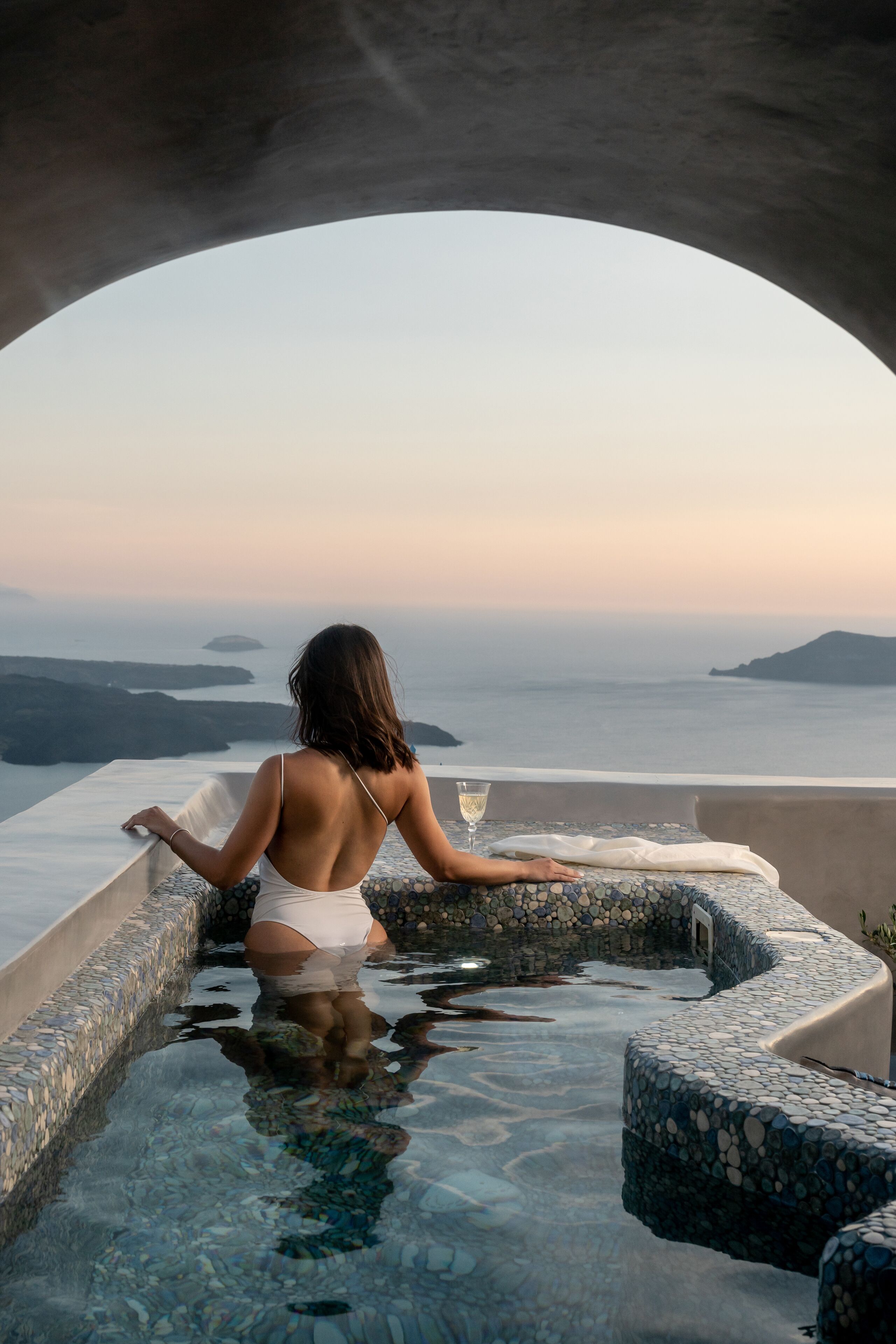 Mulher de fato de banho branco sentada em piscina infinita de pedra, com uma taça de vinho, apreciando a paisagem costeira.