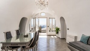 A dining area with a glass table and chairs, a chandelier, and a view of the sea through an open archway. - Luxury Cave Villa, Caldera View, Outdoor Heated Plunge Pool (Imerovigli, Thira)