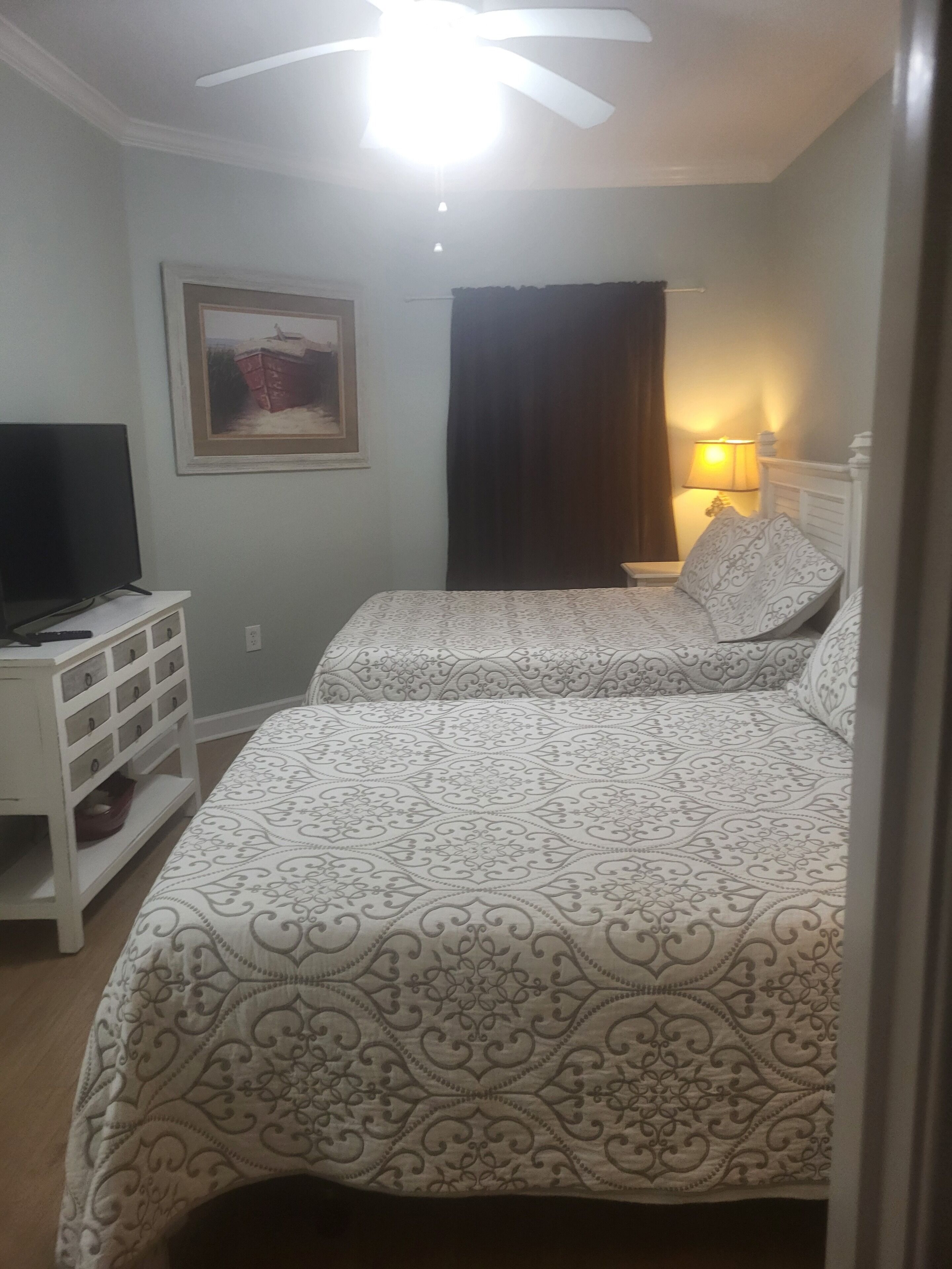 Een slaapkamer met twee bedden, een televisie op een nachtkastje, een schilderij aan de muur en een plafondventilator.