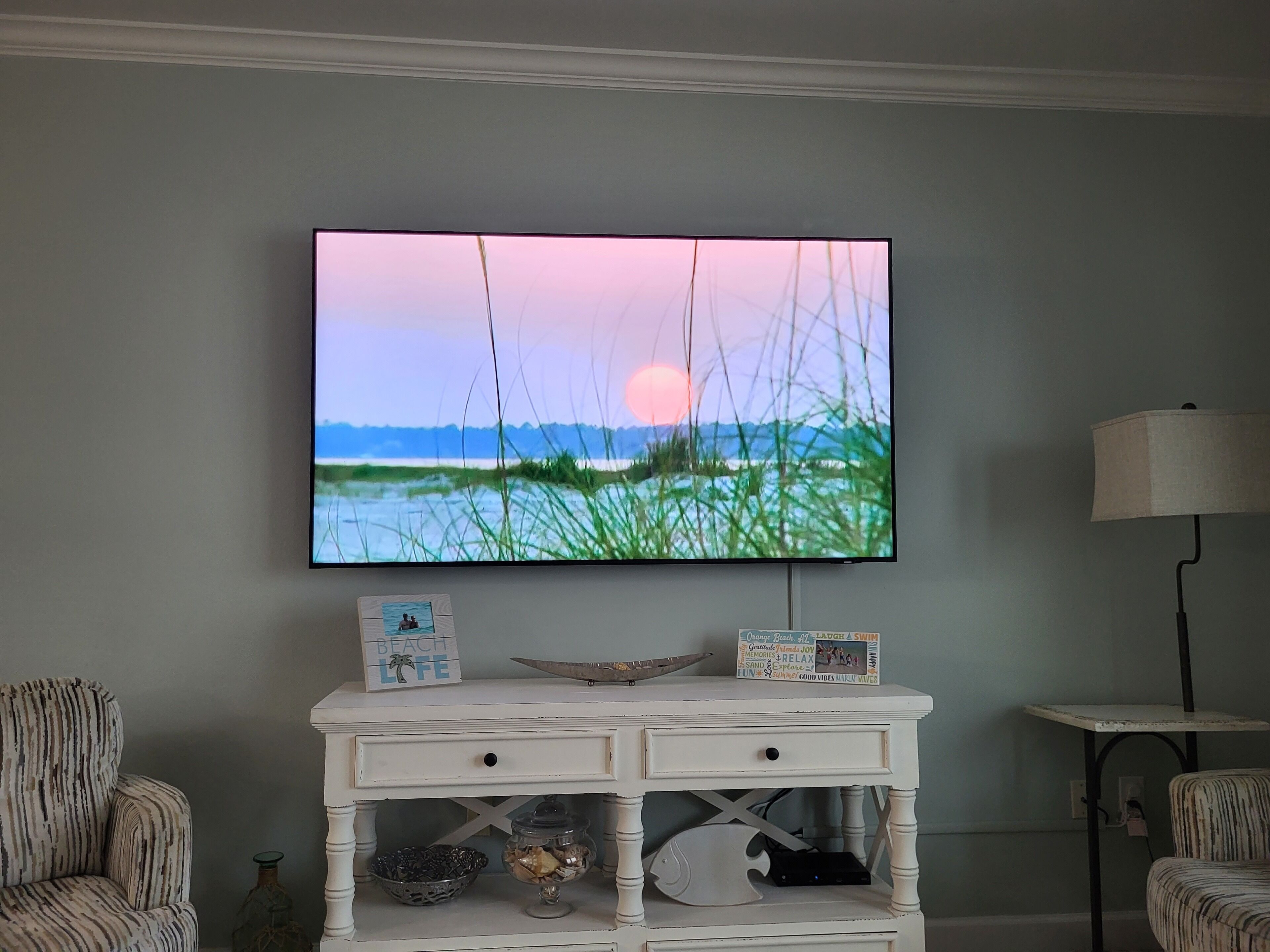 Un téléviseur fixé au mur diffuse une image de coucher de soleil sur l’eau. En dessous du téléviseur, on trouve une console blanche avec des tiroirs et des objets décoratifs.