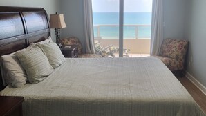Een slaapkamer met een groot bed, een plafondventilator, een nachtkastje, een lamp, een stoel en uitzicht op de oceaan door een schuifdeur.