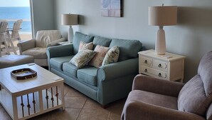 Een woonkamer met een blauwgroene bank, twee fauteuils, een salontafel en een bijzettafeltje met lampen. Een schilderij hangt aan de muur, en een raam biedt uitzicht op de oceaan.