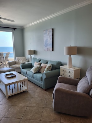 Ein Wohnzimmer mit einem türkisfarbenen Sofa, zwei Sesseln, einem Couchtisch und einem Beistelltisch mit Lampen. Ein Gemälde hängt an der Wand, und ein Fenster bietet Blick aufs Meer.