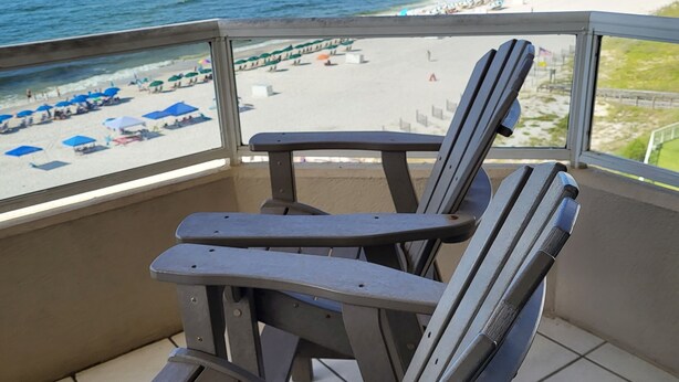Een balkon met twee Adirondack-stoelen met uitzicht op een strand met parasols en een heldere lucht.