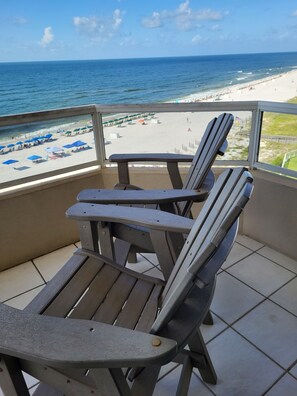 Un balcon avec deux fauteuils Adirondack donnant sur une plage parsemée de parasols, sous un ciel dégagé.