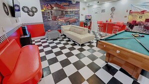 Uma mesa de bilhar com chão xadrez, um sofá e cadeiras vermelhas em um ambiente com mural de lanchonete.