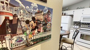 Paredes com um mural temático da Disney, com Mickey e Minnie Mouse, uma cozinha branca com micro-ondas, torradeira e fogão, e um espaço de refeições com uma cadeira e mesa.