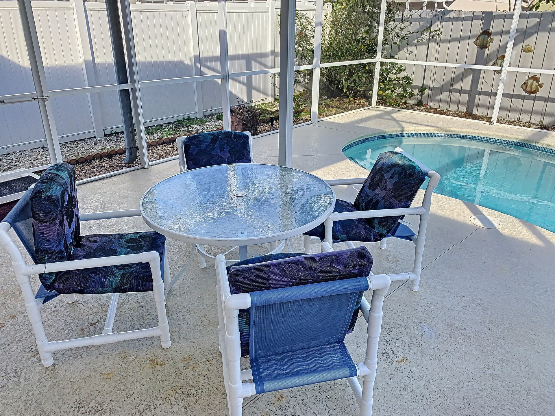 Une table ronde en verre, entourée de quatre chaises bleues à coussins, une clôture blanche et, au fond, une piscine.