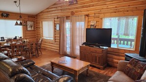 L’intérieur d’une cabane en rondins comprend un coin repas, un salon avec un canapé et un divan en cuir, un téléviseur et une cheminée.
