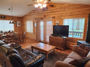 A log cabin interior with a dining area, a living room with a leather couch and a sofa, a television, and a fireplace.