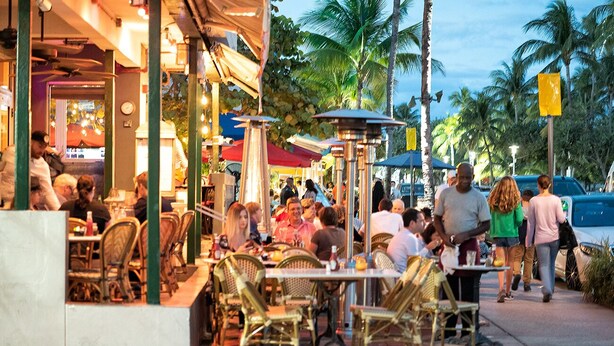 Un espace extérieur aménagé pour les repas, avec des tables et des chaises, des palmiers et une rue où circulent des voitures.