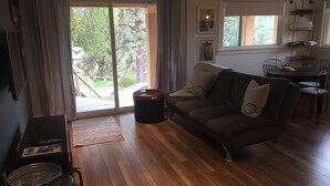A living room with a sofa, a dining table with chairs, a small black table with a tray, and a large window with curtains. - Cozy house in the shade of pines with Lake and Mountain view (Kelowna)