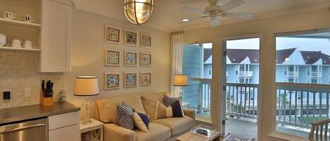 A modern living room with a sofa, coffee table, and a dining area. There are framed pictures on the wall, a ceiling fan, and large windows with a view of houses.