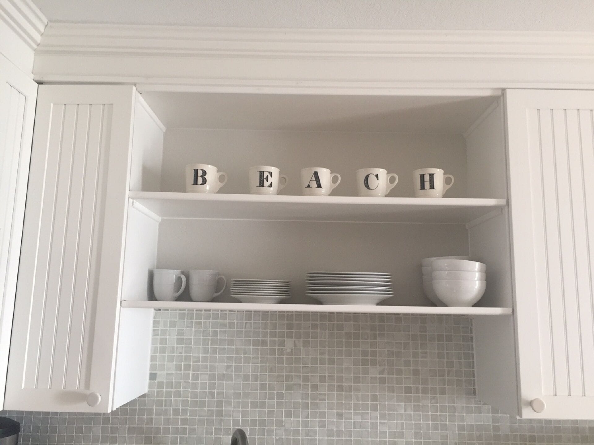 Ein Küchenschrank mit einer Ablage, auf der weiße Tassen mit dem Aufdruck 'BEACH' stehen.