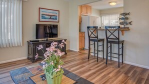 Una sala de estar con televisión, un arreglo floral y una cocina con armarios de madera y refrigerador.