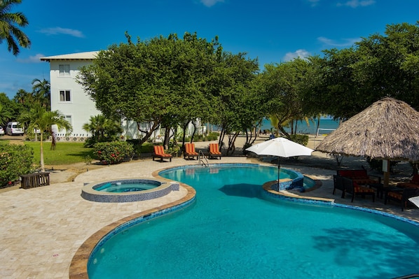 A swimming pool with lounge chairs and a thatched umbrella, surrounded by trees and a building.