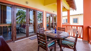 Un espacio exterior cubierto para comer con una mesa y sillas, vista a un edificio con balcón y una palmera al fondo.