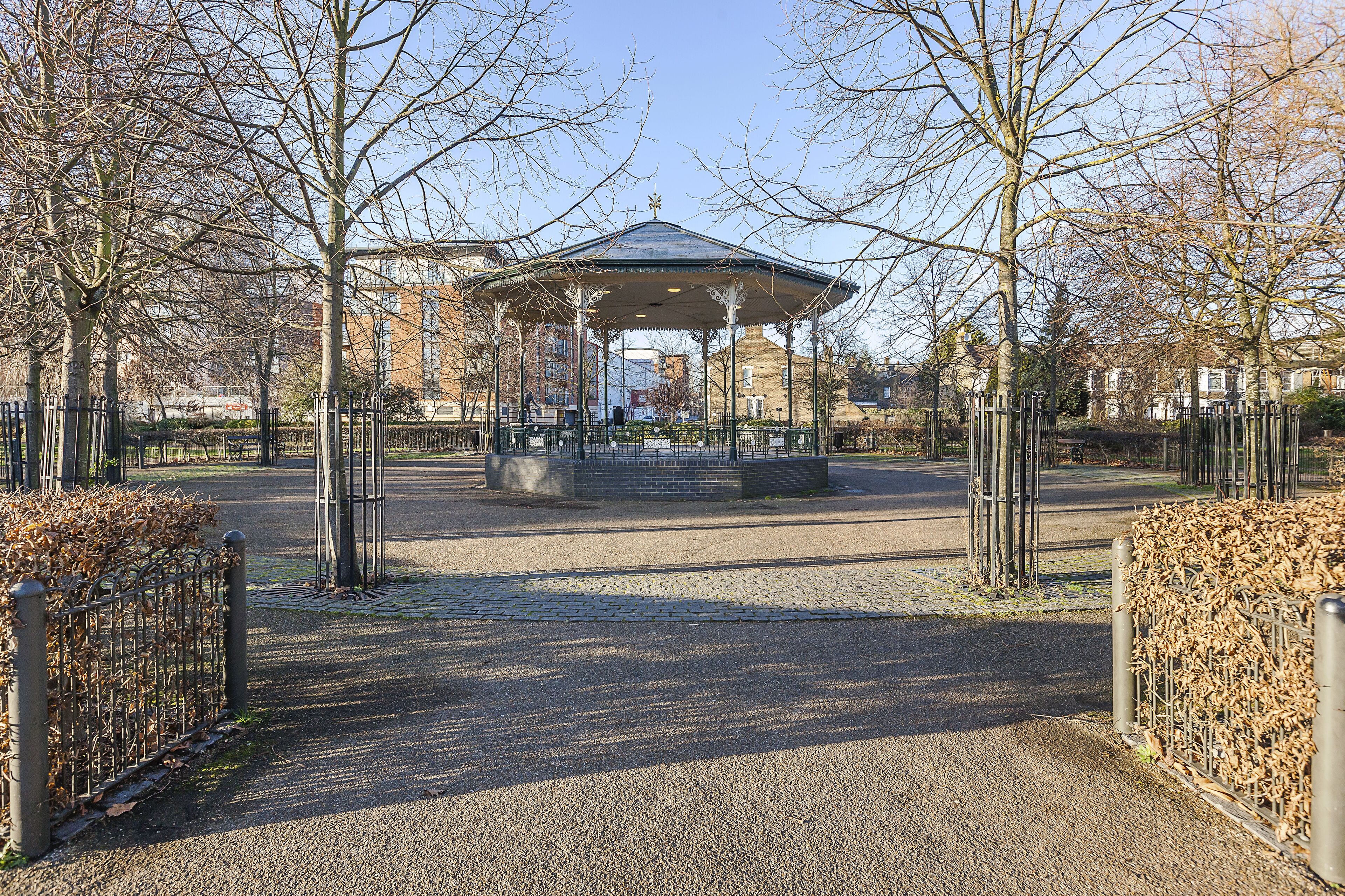 A gazebo in a park with a gravel path leading to it, surrounded by leafless trees and a metal fence.
