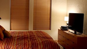 Een slaapkamer met een bed, houten zonwering, een televisie op een dressoir en een lamp.