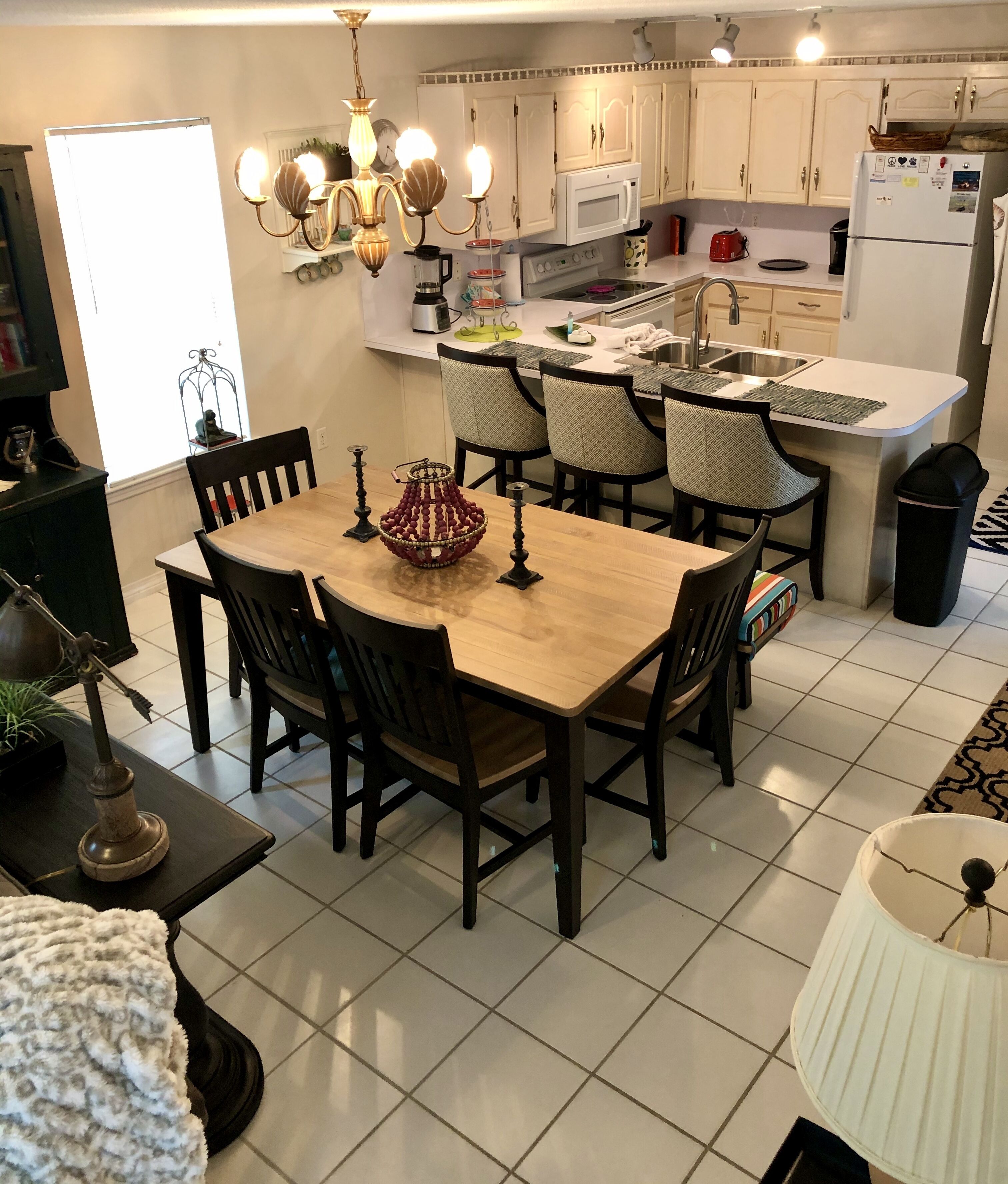 A modern kitchen with white cabinets, a central island with seating, a dining table with chairs, and a chandelier.
