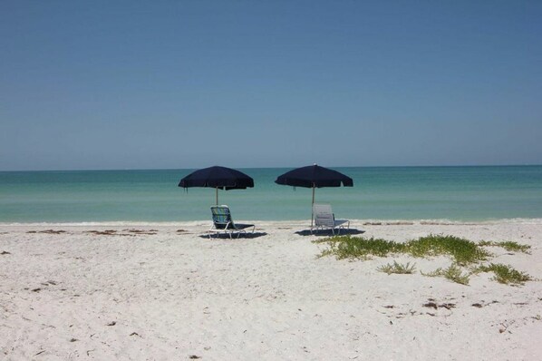 A beach with two lounge chairs and two umbrellas.